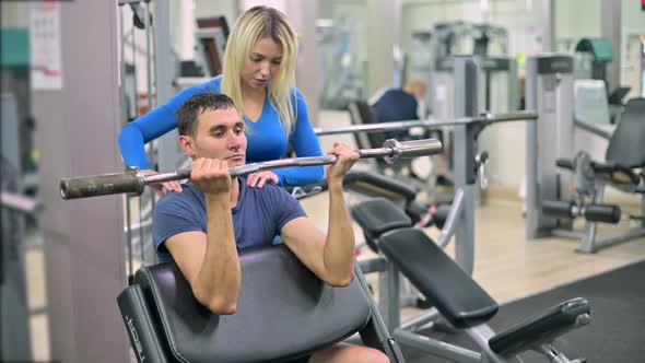 Skinny guy doing biceps exercise under the supervision of a trainer in a gym