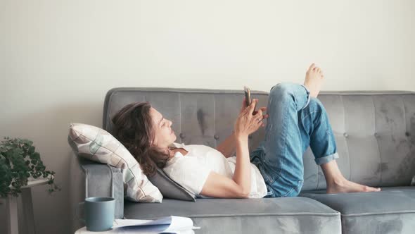 Young Cheerful Woman Lying on a Grey Couch with a Smartphone in Hands.