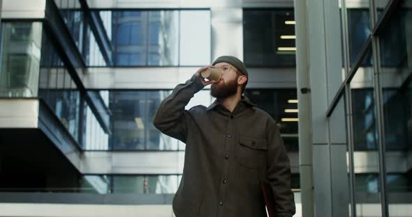 A Young Bearded Man Walks Near an Office Building and Drinks Coffee