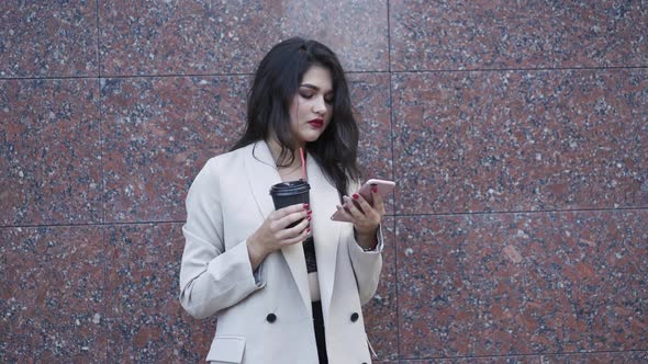 Attractive Young Girl Using Smartphone and Drinks a Cup of Beverage Outdoors