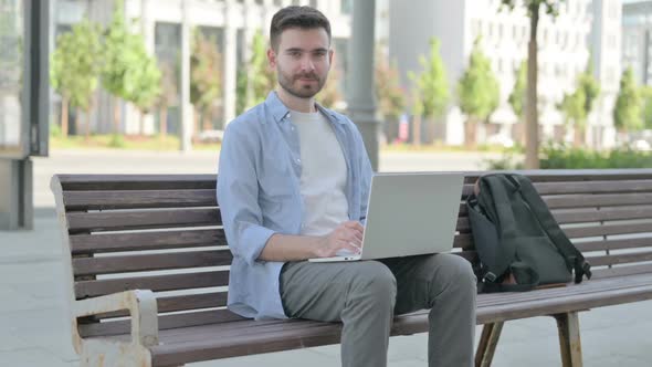 Young Man with Laptop Smiling at Camera While Sitting on Bench