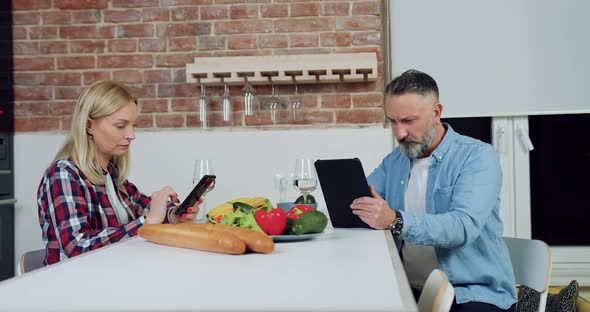 Woman Uses Phone and Sitting at Kitchen Table with Her Concentrated Bearded Husband