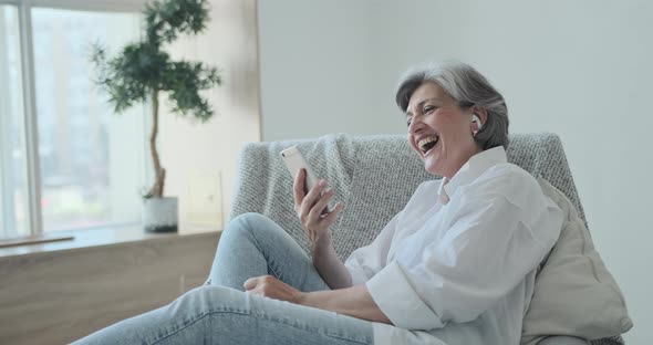 Happy Senior Old Woman in Headphones Looking Mobile Screen Enjoying Video Meeting