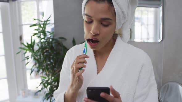 Mixed race woman brushing her teeth and using her smartphone in bathroom