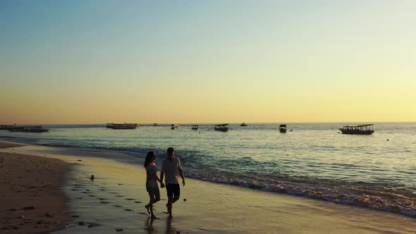 Young couple holding hands walking along exotic beach washed by sea waves, enjoying beautiful sunset