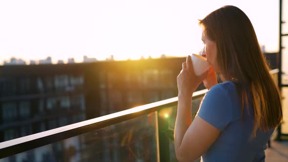 Woman with a Cup of Coffee Standing on the Balcony and Admire the Sunset