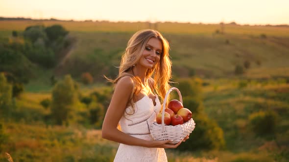 Beautiful sexy blonde girl in white dress posing in a field at sunset with a basket of fruit	