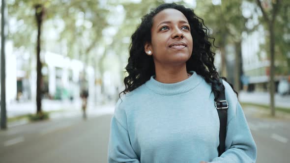 Pleased brunette African woman wearing blue sweater typing by phone and walking