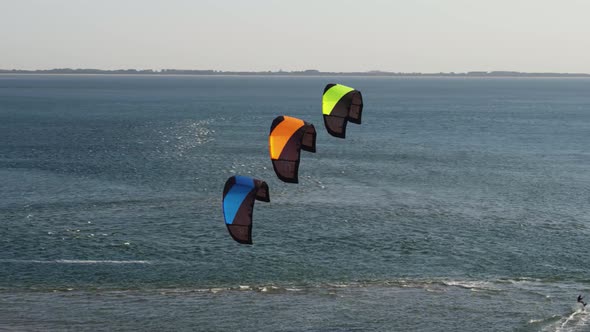 Aerial orbital reveal of a kite surfer skimming across the sea as three kites are held stationary in