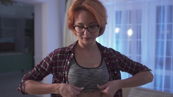 Young Attractive Woman with Short Red Hair in Glasses Puts on Sport Top at Home. Athletic Lady