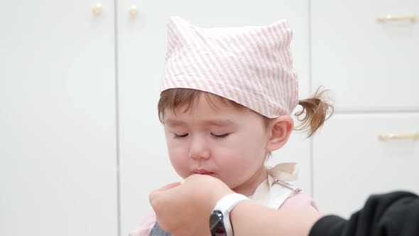 Hungry Toddler Wearing A Chef Costume In A Kids Cafe Fed By Her Mother. Close Up Shot