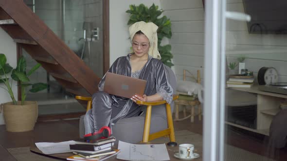 Serious Asian Young Woman in Pajamas Hair Towel and Eyeglasses Listening to Business Ideas in Laptop