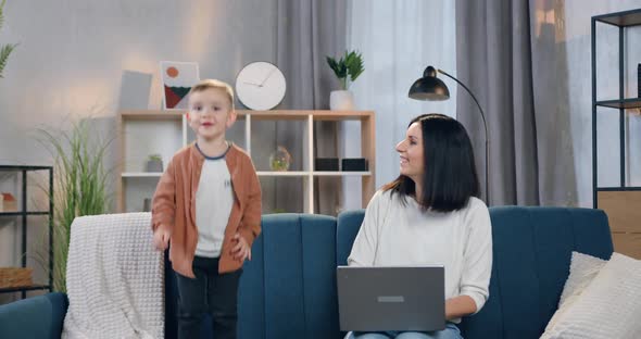Happy Mother with Black Hair Admiring of Her Cute 2-Aged Son which Jumping Near Her on Couch