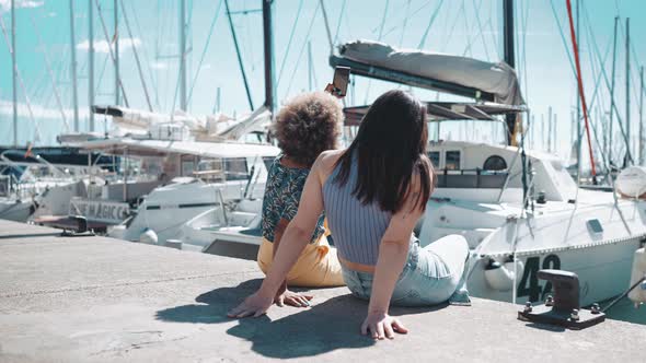 Two laughing multiethnic women sitting on the embankment near the yachts