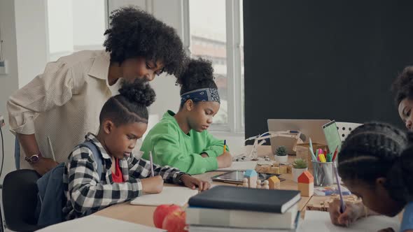 African American female teacher standing with pupils teaching writing lesson in modern classroom.