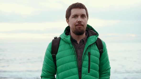 Adult Man with Beard and Backpack Is Standing Outdoor Near Sea in Cloudy Weather