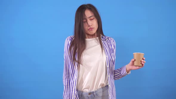Young Sleepy Asian Woman Yawns and Stands on a Blue Background with a Up of Coffee