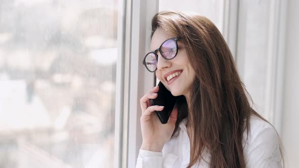 Pretty Woman in Glasses Is Making a Call Near the Window and Smiling