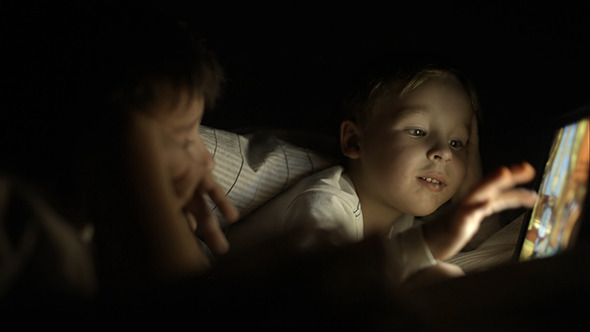 Two Boys Lying In Bed At Night And Using Pad