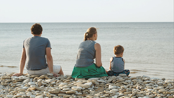 Family Of Three Sitting On Pebble Beach By Water