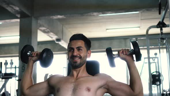 Muscular Powerful Young Man Doing Shoulders Overhead Press Lifting with Dumbbells in Fitness Gym