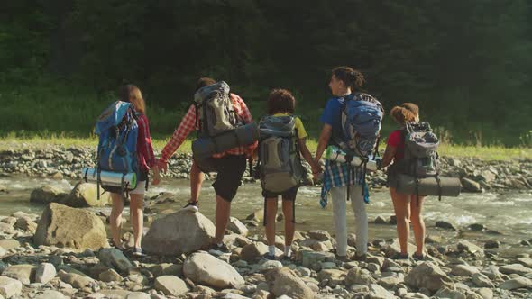 Cheerful Multicultural Backpackers Celebrating Successful Hike with Arms Raised By Mountain River