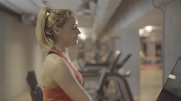 Sportive Young Caucasian Woman Drinking Water As Walking on Treadmill in Gym. Side View of Beautiful