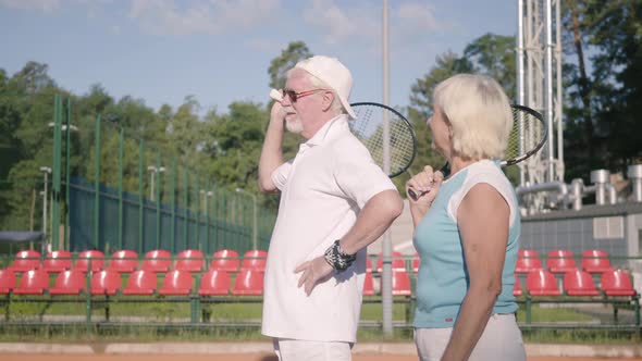 Mature Couple in Sunglasses and Tennis Rackets Standing on a Tennis Court in the Sun