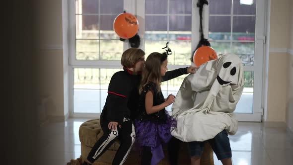 Boy and Girl Scaring Frightened Ghost on Halloween Sitting in Living Room Indoors