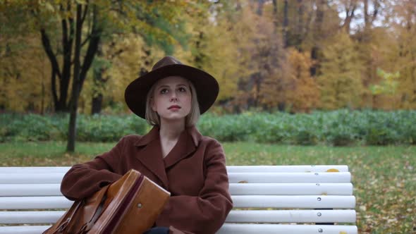 Woman in coat and hat with suitcase sits in bench in autumn park