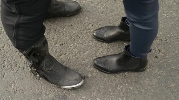 The legs of a woman in black leather boots fit a man in a motorcycle shoe