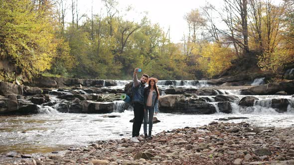 A Young Woman and a Man are Standing on the Shore of a Mountain River