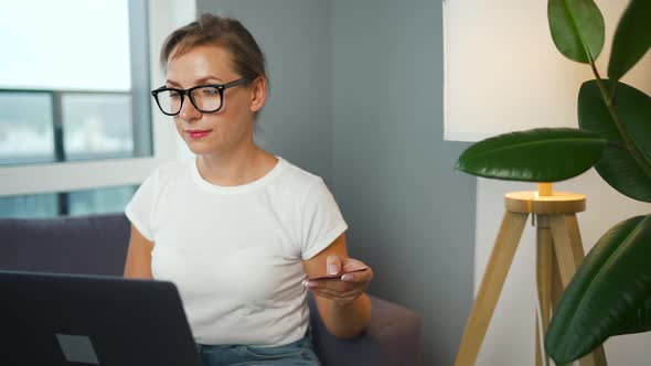 Woman with Glasses Sits on a Sofa in a Cozy Room and Makes an Online Shopping Using a Credit Card