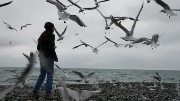 A Woman in a Hat and Jacket Feeds Gulls Flying By the Sea