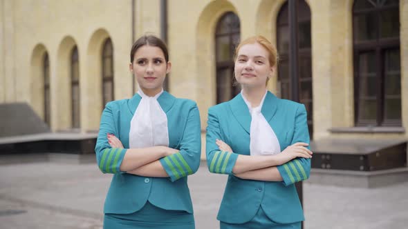 Portrait of Two Confident Young Stewardesses Standing on City Street and Crossing Hands. Positive