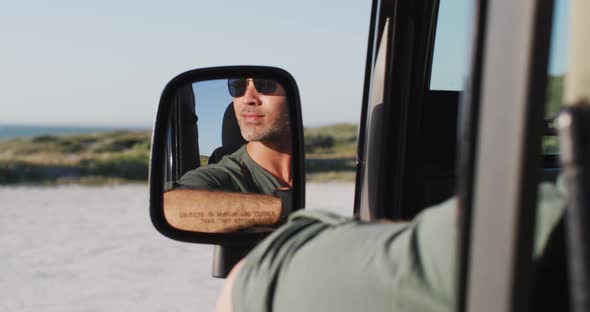 Happy caucasian man in sunglasses sitting in car reflected in side mirror on sunny day at the beach