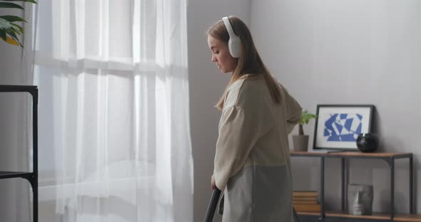 Pretty Young Woman Is Hoovering Floor in Apartment Listening To Music By Headphones and Dancing