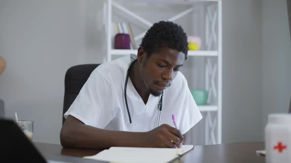 Concentrated African American Doctor Sitting at Table Writing with Pen