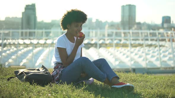 Happy Curly Haired Female Eating Fresh Apple and Sitting on Green Lawn, Health