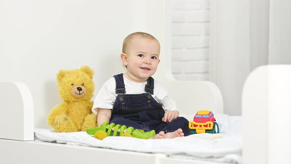 a Baby Boy in a Denim Jumpsuit Smiles and Plays with Toys While Sitting on a Bed