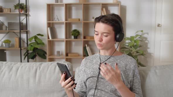 Young Woman Wear Headphones Sit on Couch at Home Communicates with Family Use Smartphone and