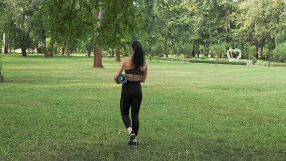 Woman in sport clothes walking with a yoga mat in city park in summer