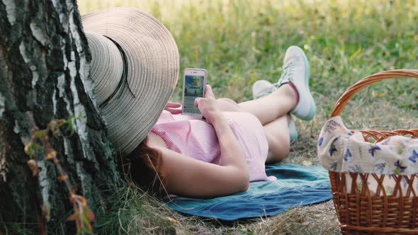 Picnic Time. Happy Young Girl with Round Hat Laying Near the Tree and Using Her Mobile Phone. Woman