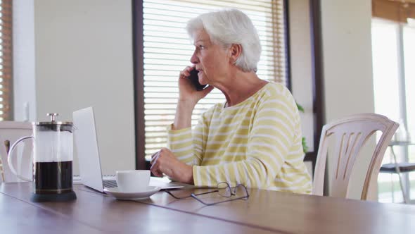 Senior caucasian woman with laptop talking on smartphone at home