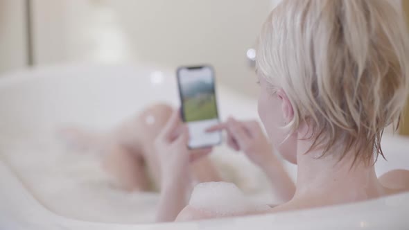 Blond Woman Scrolling Social Media While Taking Bath. Back View of Young Charming Caucasian Lady in