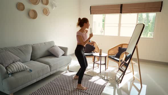 Happy Woman Dancing and Singing on the Remote Control in Front of the Mirror