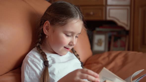 Little authentic school-age girl reads a large book while sitting on a chair at home