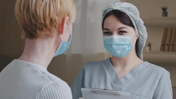 Unrecognizable Caucasian Woman Patient with Short Hair in Medical Mask Speaks to Female Doctor Nurse