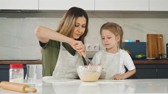 Daughter Looks at Mother Mixing Dough with Whisk at Table
