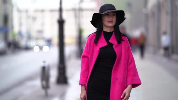 A Pretty Woman in a Pink Coat and a Black Hat Walks on a Blurry Background of a City Street. Smiles.